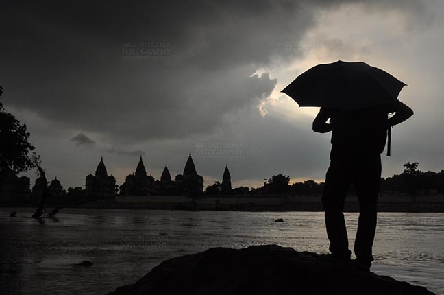 Monuments- Palaces and Temples of Orchha, Madhya Pradesh, India. (Photo: Atul Sharma) Orchha, Madhya Pradesh, India- August 20, 2012: Chhatris on the bank of river Betwa, a tourist holding umbrella enjoying cloudy- rainy season at Orchha, Madhya Pradesh, India. by Anil