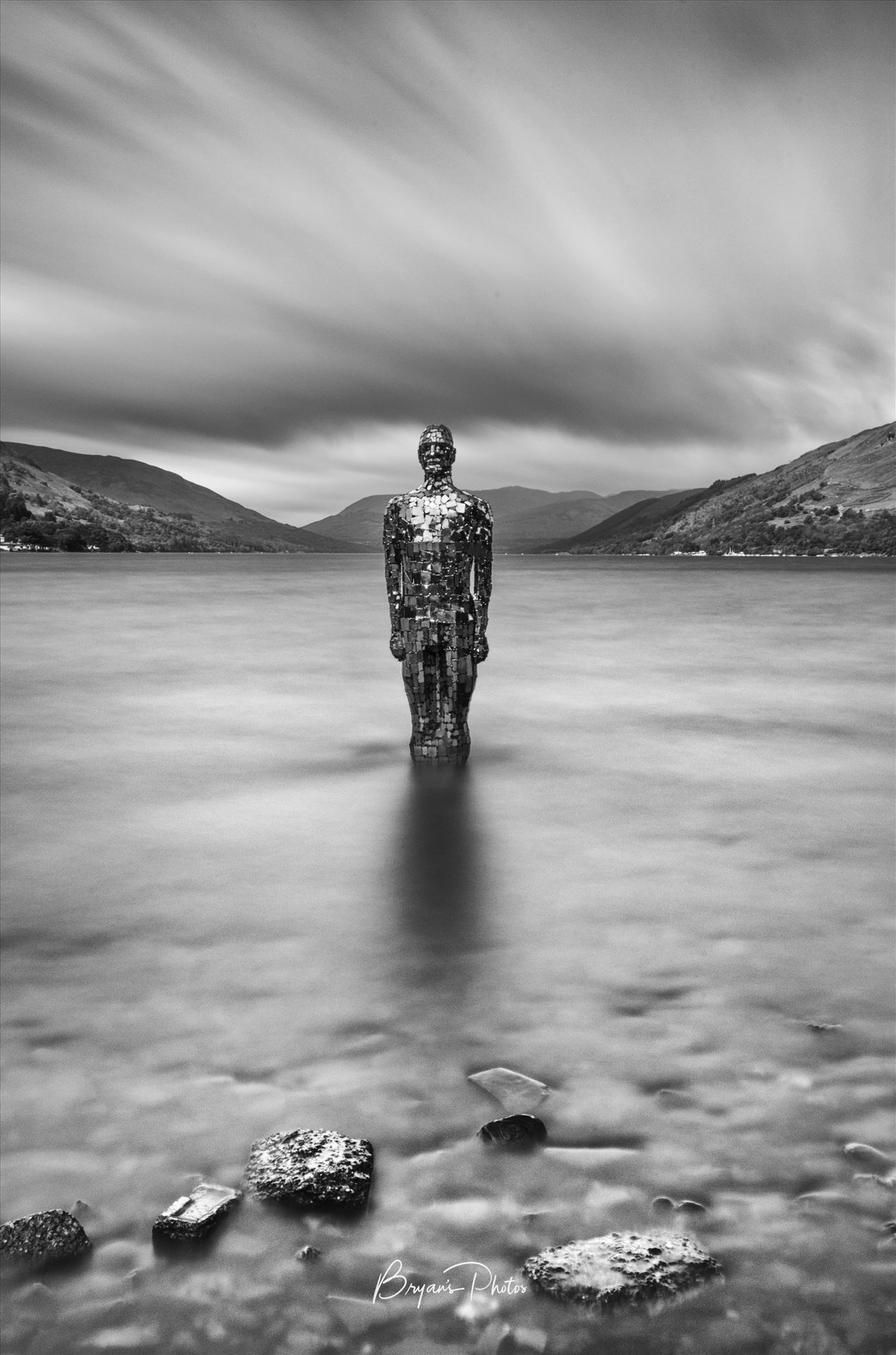 Mirror Man A black and white long exposure photograph of the still mirror man sculpture taken from St Fillans at Loch Earn. by Bryans Photos